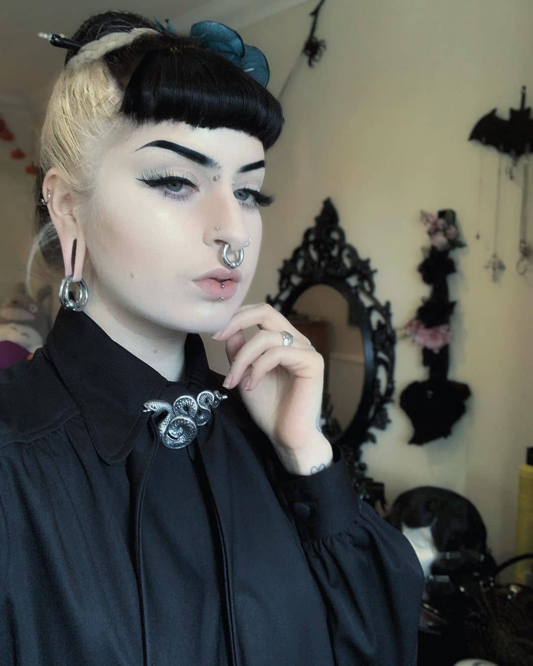 Woman with dark hair wearing a black outfit with a silver snake brooch in an interior setting.