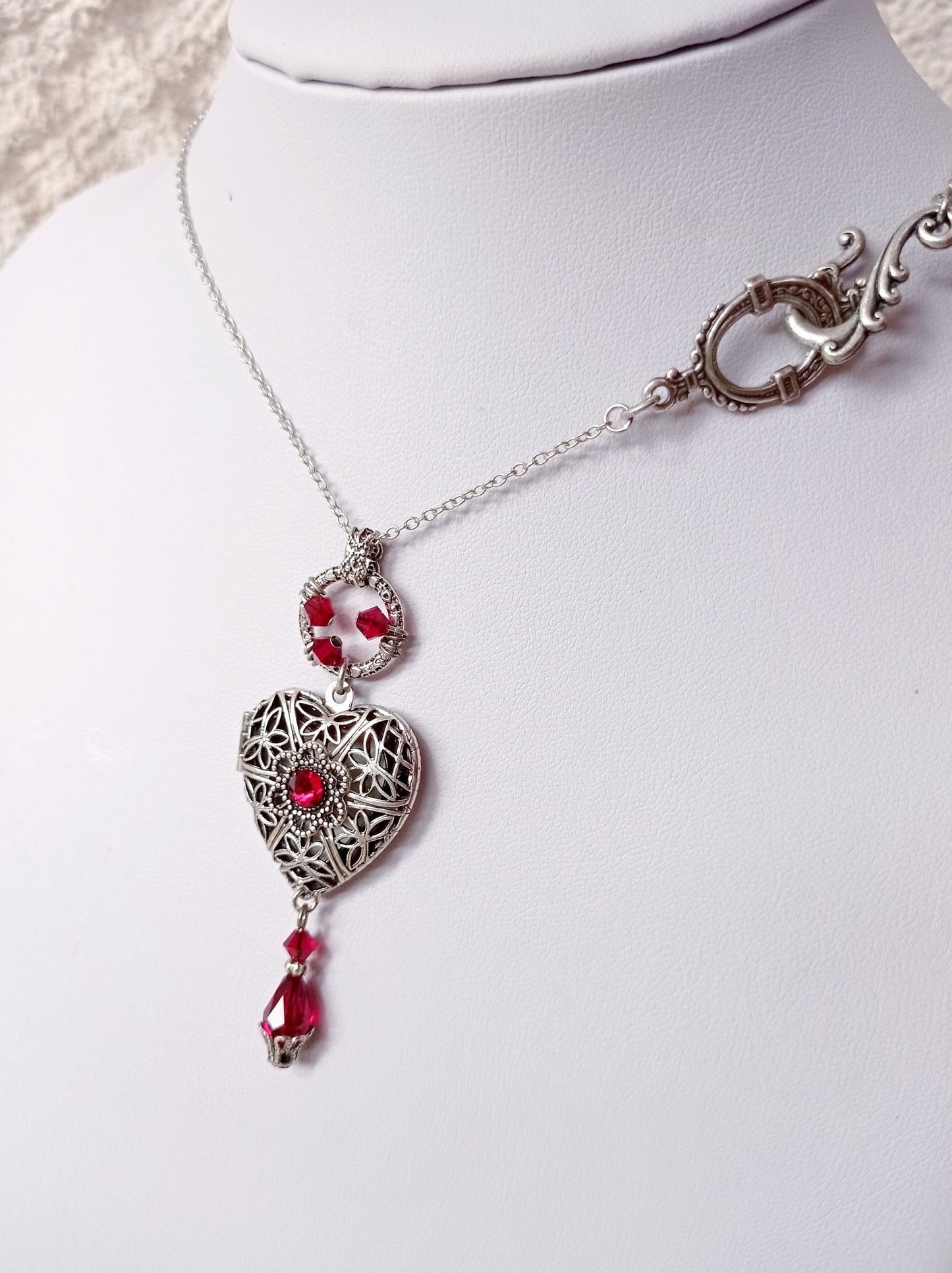 A silver necklace with an ornate front clasp features a filigree heart locket pendant. The heart has a red gem in the center and is adorned with red gemstones above and below. 