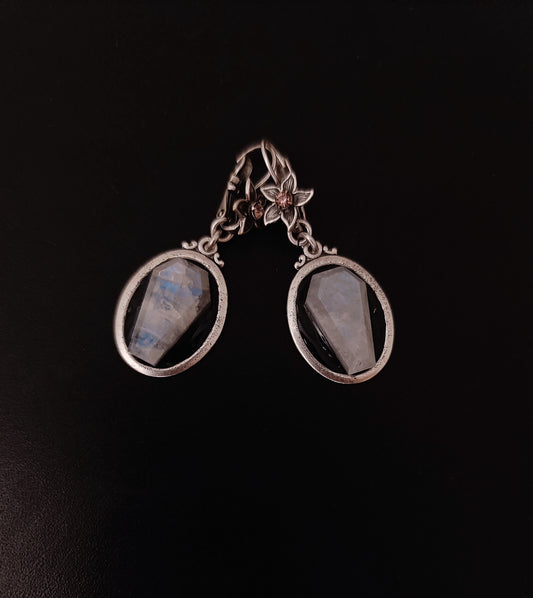 A pair of silver drop earrings with oval bezel settings holding milky white moostones with subtle blue-gray flashes, displayed on a dark black background.