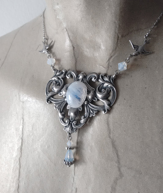 A vintage-style silver necklace featuring an ornate design, a central moonstone cabochon, and two small bird charms on either side.